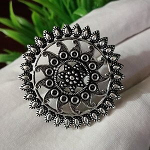 May include: A silver-colored statement ring with a detailed floral design. The ring features a central flower surrounded by a circular pattern of stylized petals and small, beaded accents. The ring is set against a white fabric background.