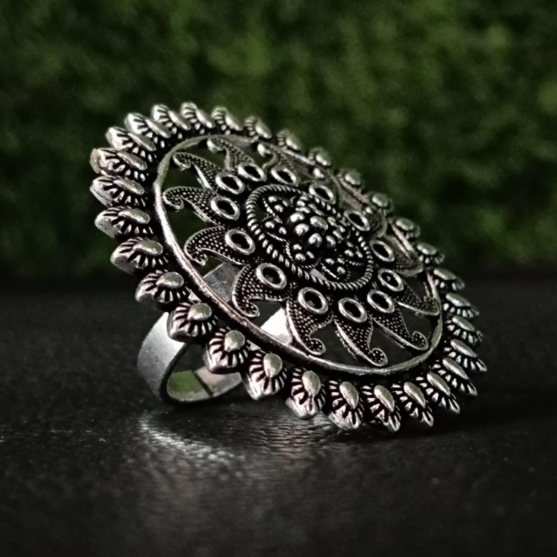 May include: A silver-toned statement ring with an intricate, floral-inspired design. The ring features a large, circular face with detailed patterns and a textured band. The ring is set against a dark background, highlighting its ornate details.