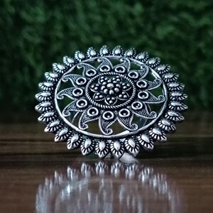 May include: A silver-colored ring with an intricate floral design. The ring features a central flower surrounded by a series of circular and petal-shaped elements. The ring's design is detailed and ornate, with a vintage or antique aesthetic.