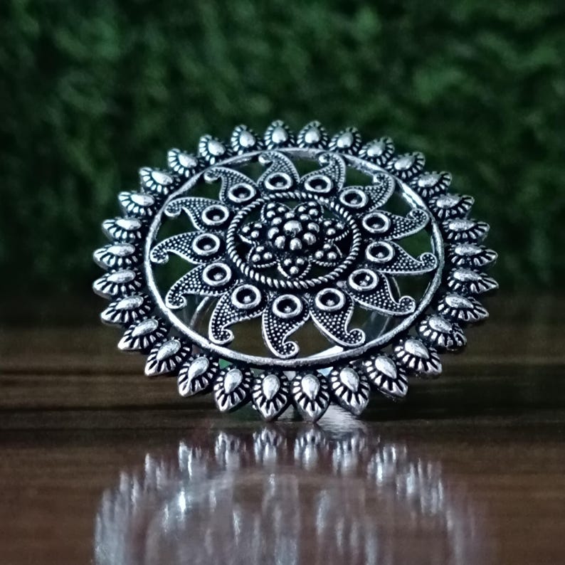 May include: A silver-colored ring with an intricate floral design. The ring features a central flower surrounded by a series of circular and petal-shaped elements. The ring's design is detailed and ornate, with a vintage or antique aesthetic.
