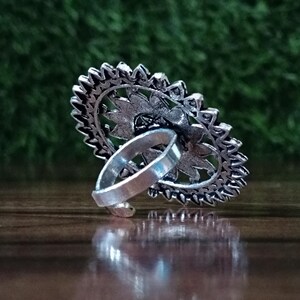 May include: A silver-toned ring with an oval, floral-patterned face. The ring's design features a detailed, openwork pattern with a central floral element and a textured border. The band is a simple, polished silver.