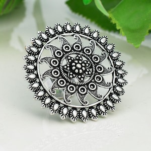 May include: A silver-colored ring with an intricate floral design. The ring features a central flower surrounded by a circular pattern of stylized petals and small, teardrop-shaped accents. The ring's design is symmetrical and detailed.
