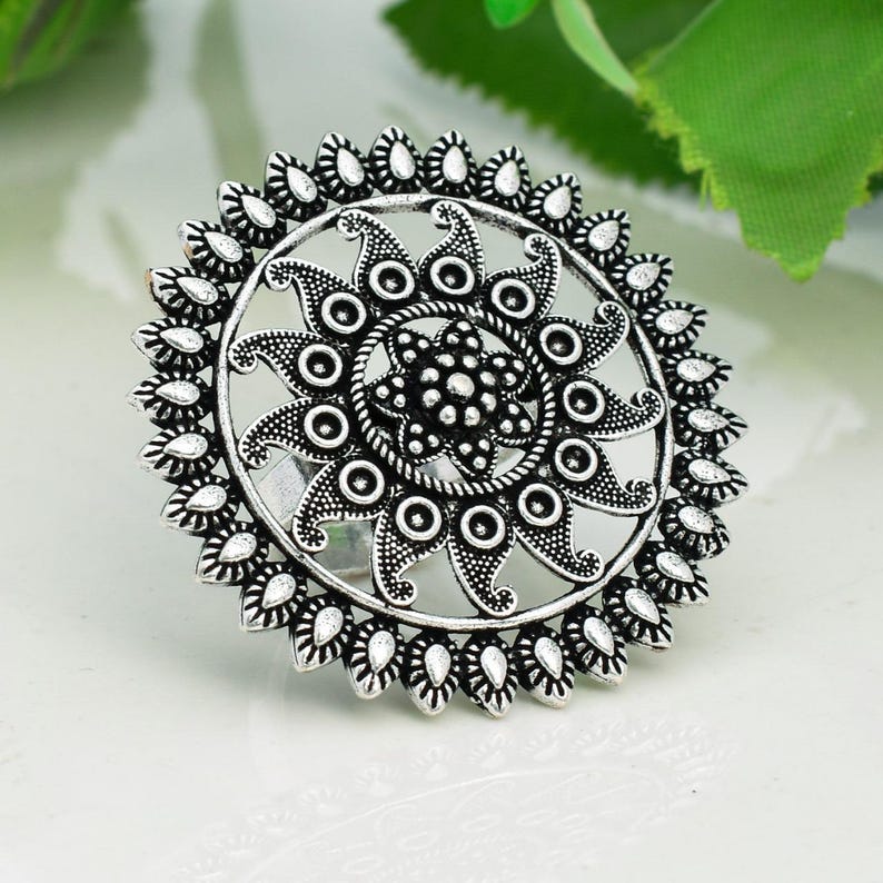 May include: A silver-colored ring with an intricate floral design. The ring features a central flower surrounded by a circular pattern of stylized petals and small, teardrop-shaped accents. The ring's design is symmetrical and detailed.