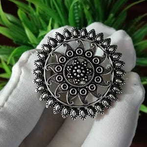May include: A silver-toned, circular ring with an intricate floral design. The ring features a central flower-like pattern surrounded by a series of smaller, petal-shaped elements. The ring is held against a white fabric background with green foliage.