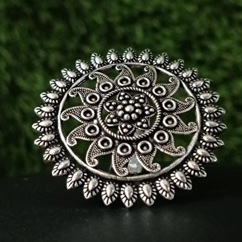 May include: A silver-colored ring with an intricate floral design. The ring features a central flower surrounded by detailed patterns and a scalloped edge. The ring is set against a dark surface with a green background.