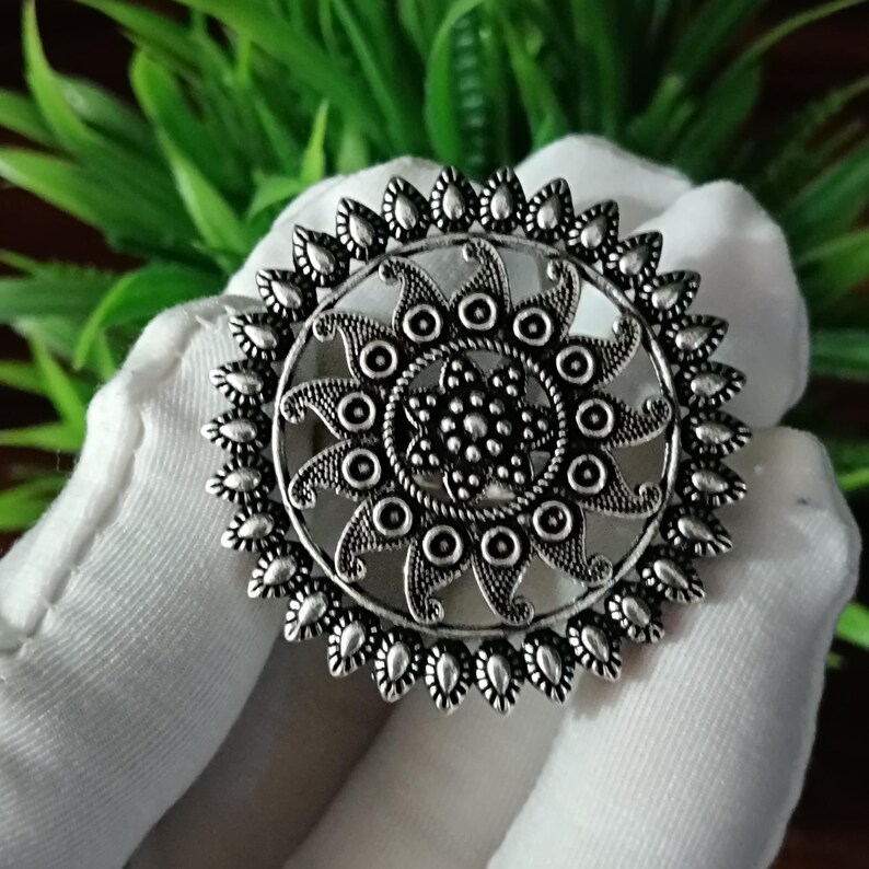 May include: A silver-colored ring with an intricate, circular design. The ring features a central floral pattern surrounded by a series of smaller circles and teardrop-shaped embellishments. The ring is held against a white background.