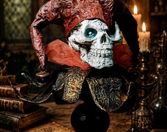 May include: A skull wearing a textured dark red jester hat with small bells, featuring bright blue eyes and a red tulle collar, mounted on a black pedestal base. The skull has a weathered white finish with visible cracks and intricate markings.