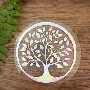 May include: A circular sticker featuring a tree of life design with a holographic finish that reflects various colors. The tree has a central trunk and spreading branches with numerous leaves, all enclosed within a thin circular border.