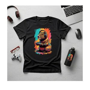 May include: Black t-shirt with a colourful illustration of a capybara wearing headphones and holding a game controller. The design includes a vibrant background with splashes of teal, orange, and pink. A black water bottle with a matching graphic is also visible.