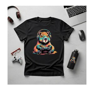 Puede incluir: Camiseta negra con una ilustración colorida de un carpincho con auriculares y un mando de videojuegos. También se ve una botella de agua a juego y otros accesorios de juego.