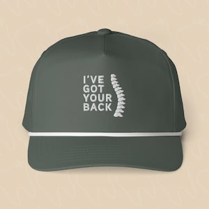 Puede incluir: Una gorra de béisbol gris oscuro con el texto bordado en blanco I'VE GOT YOUR BACK junto a un gráfico bordado en blanco de una columna vertebral humana. La gorra incluye un detalle de cordón blanco horizontal en la parte frontal.