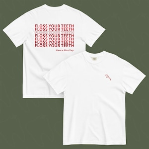 May include: Two white t-shirts. One shirt has the text "FLOSS YOUR TEETH" repeated in red on the back and "Have a Nice Day" below. The other shirt has a red graphic on the front. Both shirts are short-sleeved.