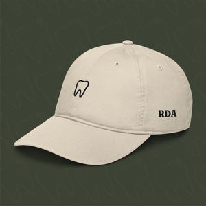 Könnte beinhalten: Eine beige Baseballkappe mit einer schwarzen Umrandung eines Zahns und den Buchstaben RDA, die auf der Seite gestickt sind.
