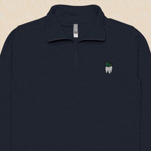 May include: Navy blue quarter-zip pullover with a small embroidered tooth and plant design on the chest. The long-sleeved shirt has a ribbed texture and a stand-up collar. The zipper is a dark metal colour.