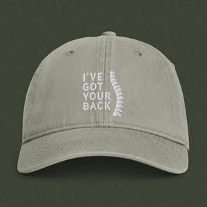 Könnte beinhalten: Eine salbeigrüne Baseballkappe mit weißem Sticktext I'VE GOT YOUR BACK, ergänzt durch eine weiße Stickgrafik einer menschlichen Wirbelsäule, die vertikal rechts neben dem Text auf dem vorderen Teil der Kappe platziert ist.