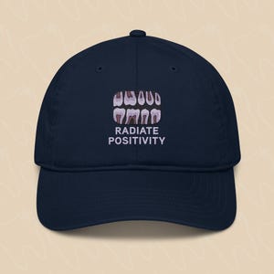 May include: Navy blue baseball cap with an embroidered design of teeth and the text "RADIATE POSITIVITY".