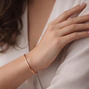 Könnte beinhalten: Ein zartes Armreif-Armband aus Roségold. Das dünne, gebogene Armband wird am Handgelenk getragen, mit einem weichen, neutralen Hintergrund. Das Armband ist ein schlichtes, elegantes Schmuckstück.