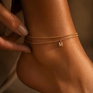 May include: A double-layered gold chain anklet featuring a small, dangling letter M charm. The delicate jewelry piece consists of two thin, interconnected metallic chains that wrap around the ankle, with the letter pendant centered on the lower chain.