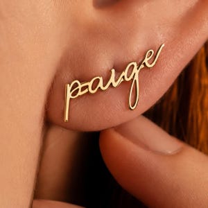 May include: A gold-colored earring in the shape of the name "Paige" in a cursive script. The earring is worn on an ear, with the word extending horizontally across the earlobe. The design is simple and elegant, with a focus on the personalized name.