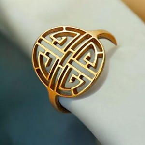 Puede incluir: Un anillo de color dorado con un diseño calado. El anillo presenta una pieza central circular con un intrincado patrón geométrico. La banda y el diseño circular están hechos del mismo metal dorado. El anillo se muestra sobre una superficie blanca.