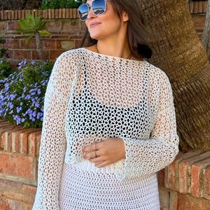 Puede incluir: Un top de crochet blanco con mangas largas y una falda de crochet blanca a juego. La parte superior es de tejido abierto, revelando una parte superior negra debajo. La falda tiene el texto "lou lou". El atuendo se usa al aire libre.