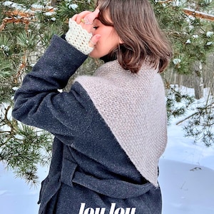 Puede incluir: Una mujer con un abrigo gris y un chal de punto beige. La imagen está ambientada en un entorno nevado al aire libre con árboles de hoja perenne al fondo. El texto "lou lou studio" está en la parte inferior.