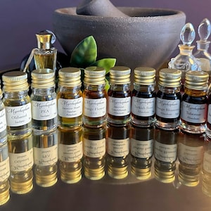May include: A selection of small glass bottles with gold lids, containing amber and dark liquids. Each bottle is labelled with its contents. The bottles are arranged on a reflective surface, with a mortar and pestle in the background. The bottles are for essential oils.