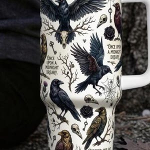 May include: A white stainless steel tumbler with a handle and lid, featuring black and brown raven illustrations, skulls, keys, spiderwebs, and roses. The text reads: Quoth the raven nevermore, Once upon a midnight dreary, and Perched and sat and nothing more.