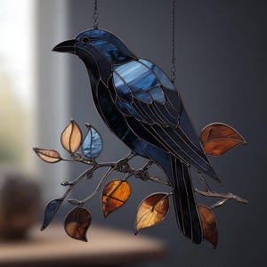 Puede incluir: Vidriera de un cuervo posado en una rama con hojas coloridas. El ave es principalmente azul oscuro con acentos azules más claros, y las hojas son tonos de naranja, marrón y azul. La obra de arte está suspendida por una cadena.