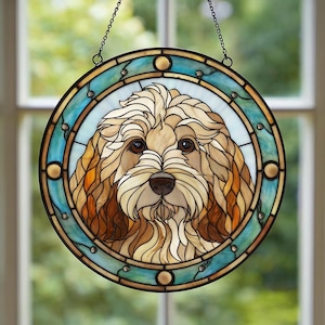 Puede incluir: Vidriera redonda que representa la cara de un perro. El perro tiene pelaje marrón claro y crema, con ojos y nariz marrones. La ventana tiene un borde turquesa y dorado y está suspendida por una cadena.