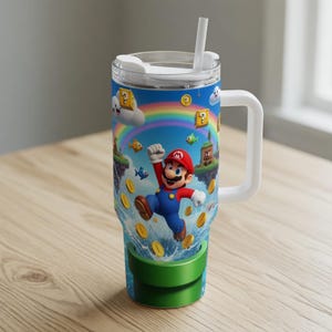 May include: A large, insulated tumbler featuring a vibrant design of the video game character Mario jumping in front of a rainbow. The tumbler has a white handle, lid, and straw. The design includes coins, fish, and floating islands.