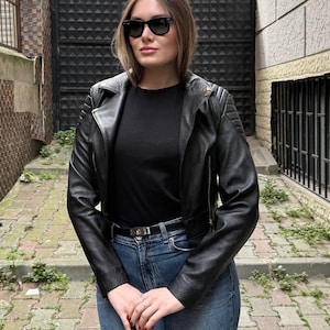 Puede incluir: Chaqueta de cuero negra con detalles acolchados en los hombros, combinada con una camiseta negra y vaqueros azules. La chaqueta tiene un estilo motero clásico con cierre de cremallera. La persona lleva gafas de sol.