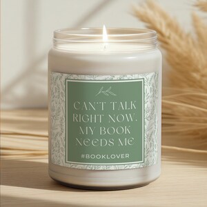 Pode incluir: Uma vela branca acesa num frasco de vidro transparente. O rótulo verde apresenta o texto branco "CAN'T TALK RIGHT NOW. MY BOOK NEEDS ME" e a hashtag #BOOKLOVER. A vela está numa superfície de madeira.