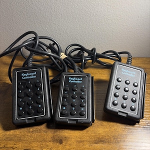 May include: Three black rectangular keyboard controllers with attached black cords. Each device features a numeric keypad with twelve circular buttons labeled 0 through 9, an asterisk, and a pound sign, with the text Keyboard Controller printed in blue.