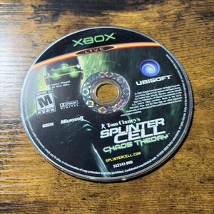 May include: An Xbox game disc for Tom Clancy's Splinter Cell Chaos Theory. The disc features a dark green graphic of a character with glowing goggles, the Xbox Live logo, an ESRB Mature 17+ rating, Ubisoft and Microsoft logos, and text.