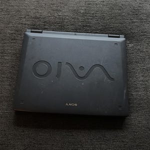 May include: A dark gray Sony VAIO laptop with a closed lid. The top surface features the large, embossed VAIO logo in the center and the Sony brand name printed in smaller white letters near the bottom edge, with visible surface scuffs.