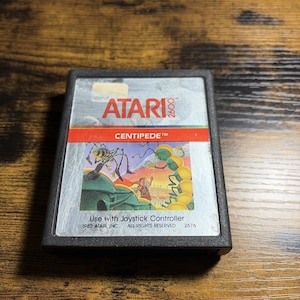 Peut inclure: Une cartouche de jeu vidéo Atari 2600 noire pour le jeu Centipede. L'étiquette présente le logo Atari, le titre Centipede en rouge et une illustration colorée d'un personnage face à un gros insecte. Le texte inclut Use with Joystick Controller.
