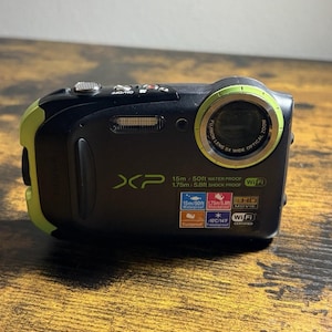 May include: A black and lime green digital camera with a Fujinon lens. The front features the text XP, 15m/50ft waterproof, 1.75m/5.8ft shockproof, and Wi-Fi. Icons indicate features including waterproof, shockproof, full HD movie, dustproof, and freezeproof.