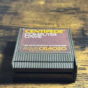 May include: A black Atari Centipede computer game cartridge with a rectangular label. The label features yellow and white text reading CENTIPEDE COMPUTER GAME, USE WITH JOYSTICK CONTROLLERS, ATARI CXL4020, and 1982 Atari, Inc. All Rights Reserved.