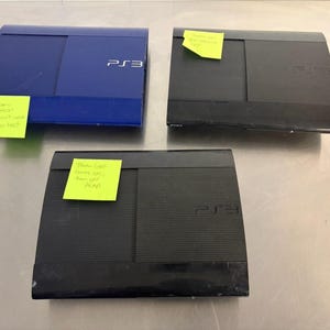 May include: Three PlayStation 3 game consoles, one blue and two black, each featuring a ribbed top surface and the PS3 logo. Each console has a yellow sticky note attached with handwritten text describing various power and display status issues.