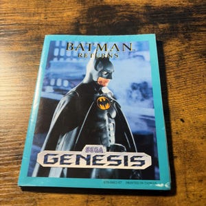 May include: A rectangular Batman Returns video game instruction manual for the Sega Genesis console. The cover features a graphic of Batman in a black suit with a yellow bat emblem, the text Batman Returns, the Sega Genesis logo, and 670-0483-07.