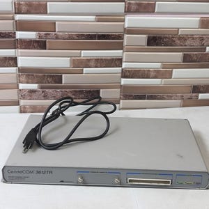 May include: A grey CentreCOM 3612TR network switch featuring a black power cord, a front-facing firmware cassette slot, an interrepeater bus port, and status indicator lights. The device has a flat, rectangular metal chassis design.