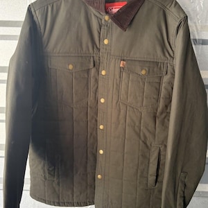 May include: An olive green quilted jacket featuring a brown corduroy collar, a front button closure with gold-toned snaps, two chest pockets with flaps, and two side pockets. The interior label displays the Coleman brand name in white text.