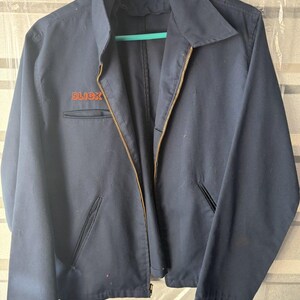 May include: A navy blue work jacket featuring a pointed collar, a full-length front zipper, and two slanted waist pockets. The word SLICK is embroidered in orange thread on the right chest. A white label with the text Workrite is at the neck.
