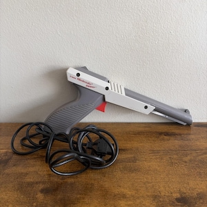 May include: A gray and white Nintendo Zapper light gun controller featuring a red trigger and a long barrel. The device includes a black coiled cord with a connector plug attached to the base of the handle, displaying the Nintendo Zapper logo.