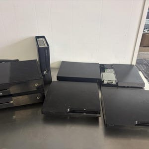 Op de afbeelding: Een verzameling zwarte videogameconsoles, waaronder een Xbox One, een Xbox 360, een PlayStation 4 met een open harde schijf-bay en twee PlayStation 3-units. De consoles zijn gerangschikt op een plat oppervlak en tonen diverse hardwareontwerpen.