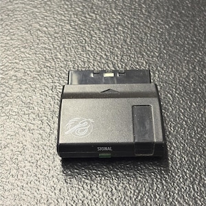 May include: A black rectangular electronic adapter featuring a multi-pin connector at the top, a small upward-pointing arrow, a circular logo on the left side, and the word SIGNAL printed in white text above a small green indicator light.