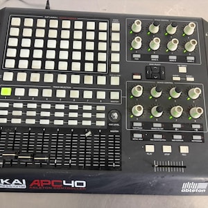 Puede incluir: Un controlador Ableton Akai Professional APC40 en color negro, con una cuadrícula de botones cuadrados, múltiples perillas giratorias con luces indicadoras verdes, botones de selección de pista y faders verticales para mezcla.
