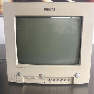 Könnte beinhalten: Ein cremefarbener Philips CRT-Monitor im Vintage-Stil mit dunklem Bildschirm. Der Monitor hat eine rechteckige Form mit abgerundeten Ecken. Er verfügt über mehrere Tasten und Bedienelemente an der Unterseite, darunter "action" und nummerierte Tasten. Das Philips-Logo ist oben sichtbar.