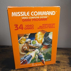May include: An orange Atari Missile Command video game box featuring the title, 34 video games, and text about interplanetary ballistic missiles. The central illustration depicts a person in a flight suit operating a console with missiles.
